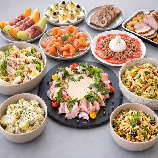 A styled spread of multiple prepared dishes on plates and bowls, including sliced cured meats, eggs, salad, pasta, and a central platter with sliced meats, greens, and a cream-colored center, arranged on a light gray surface.